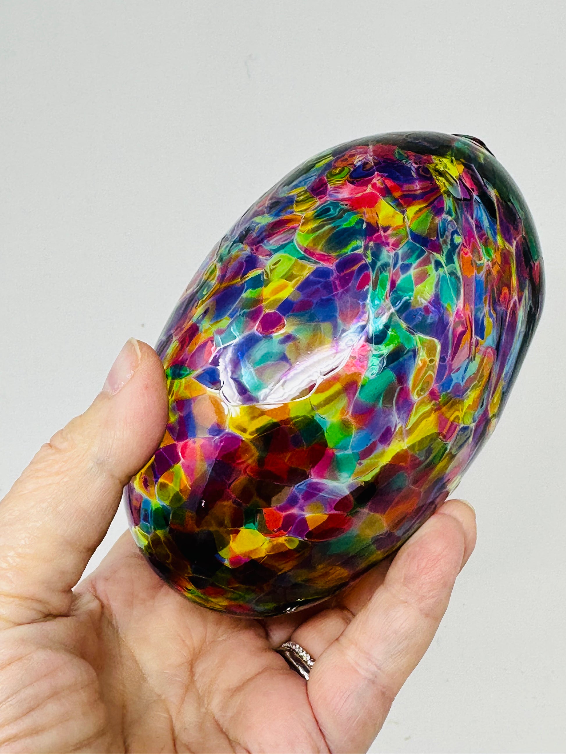 A close-up of a multicolored Chuppah glass designed by Marcela Rosemberg resting in a hand, designed for the glass-breaking ritual where it is smashed at a Jewish wedding