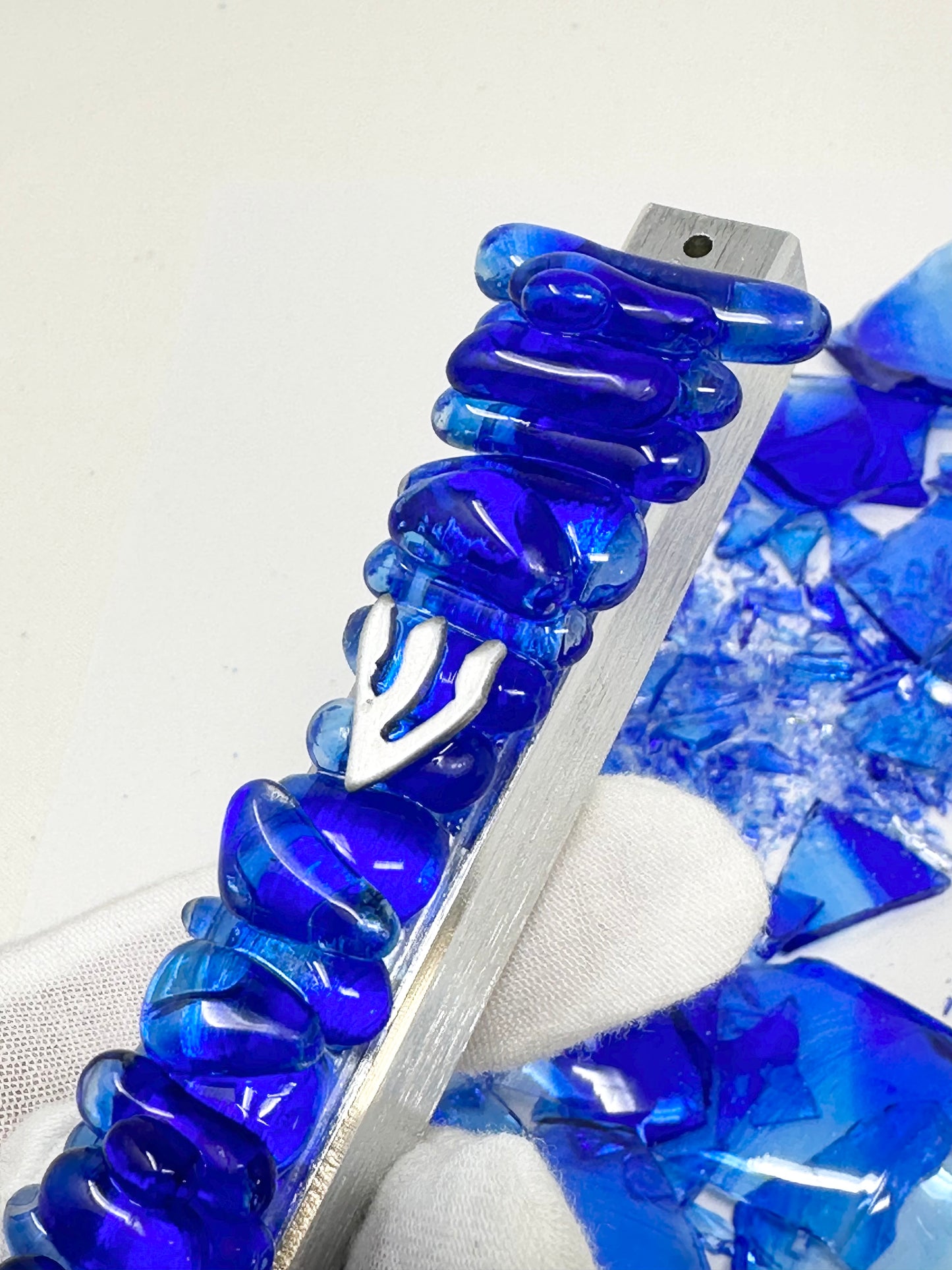 A wedding mezuzah in vibrant cobalt blue, with leftover Chuppah glass shards in the background, showcasing the transformation of the shards into a pearl-like jewel design.