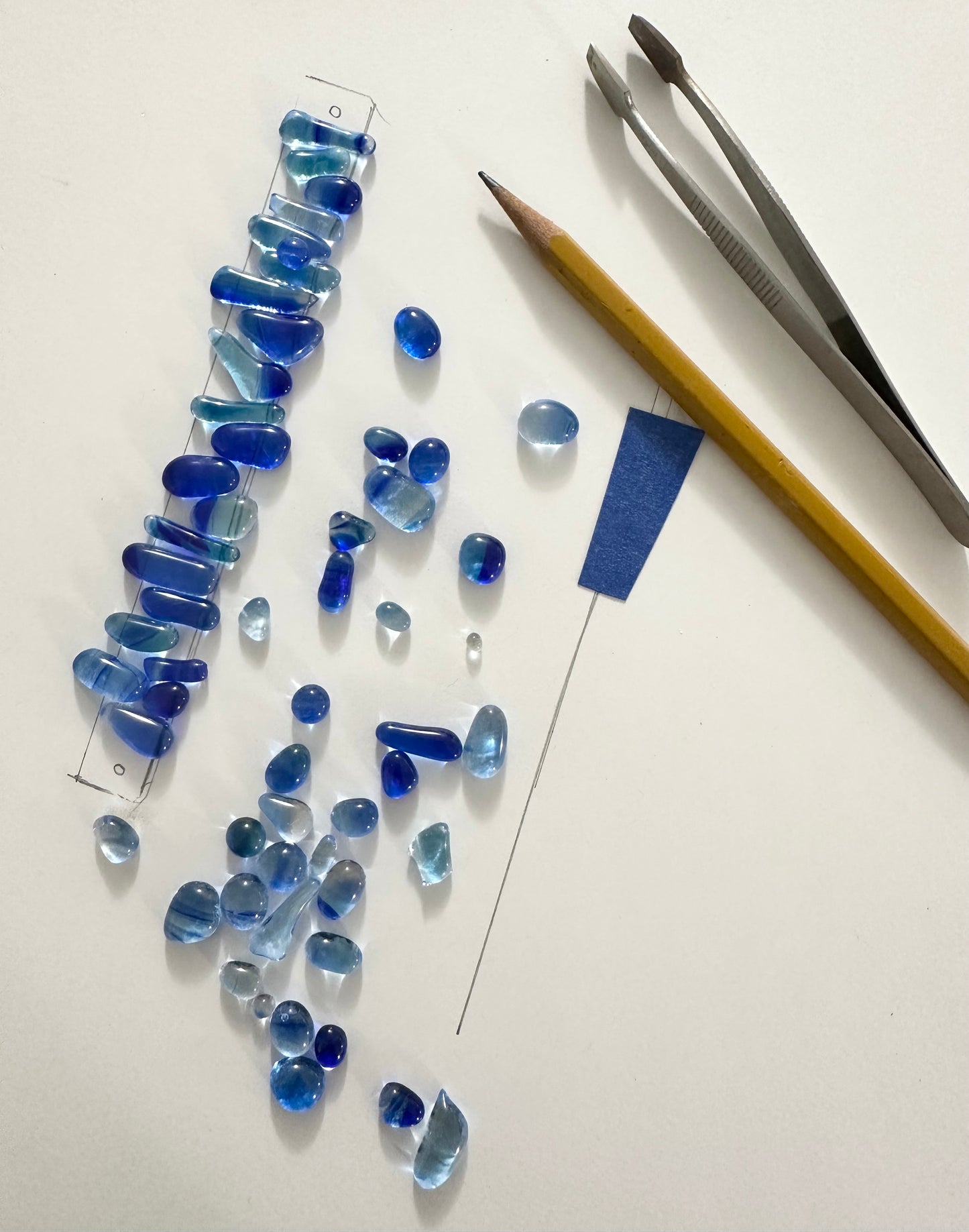 Pearl-like jewels arranged on a white background, alongside a pair of tweezers and a pencil, tools used for crafting intricate designs.