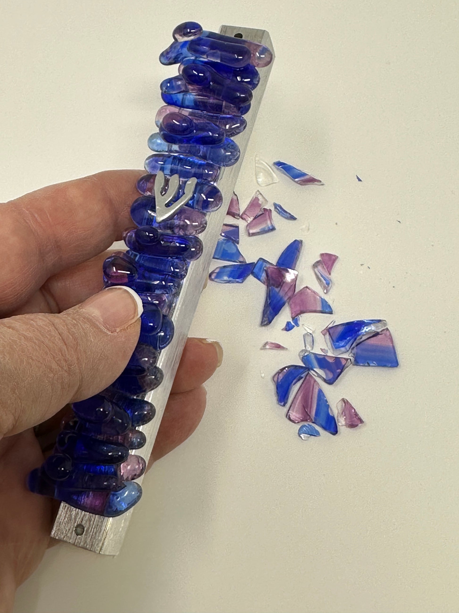 A wedding mezuzah in vibrant cobalt blue and purple, held in a hand, with leftover Chuppah glass shards in the background, highlighting the transformation of the shards into a pearl-like jewel design.