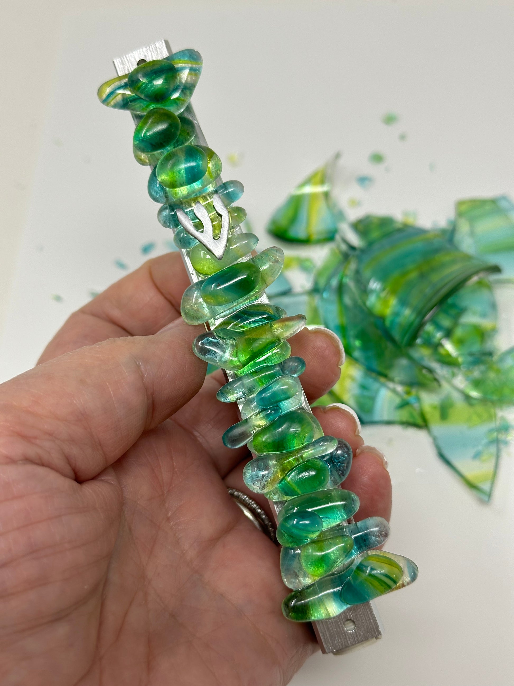 A wedding mezuzah in green and yellow, held in a hand, with leftover Chuppah glass shards in the background, highlighting the transformation of the shards into a pearl-like jewel design.