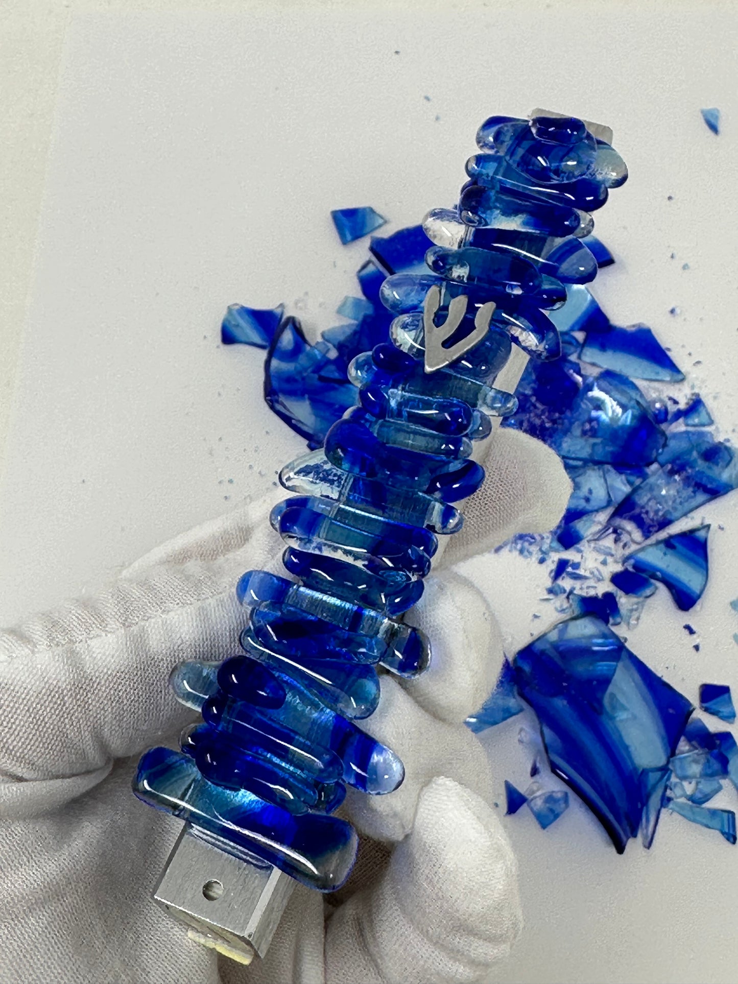 A blue wedding mezuzah held in a hand, resting on a white glove, with leftover Chuppah glass shards visible in the background, highlighting the crafting process and final piece.