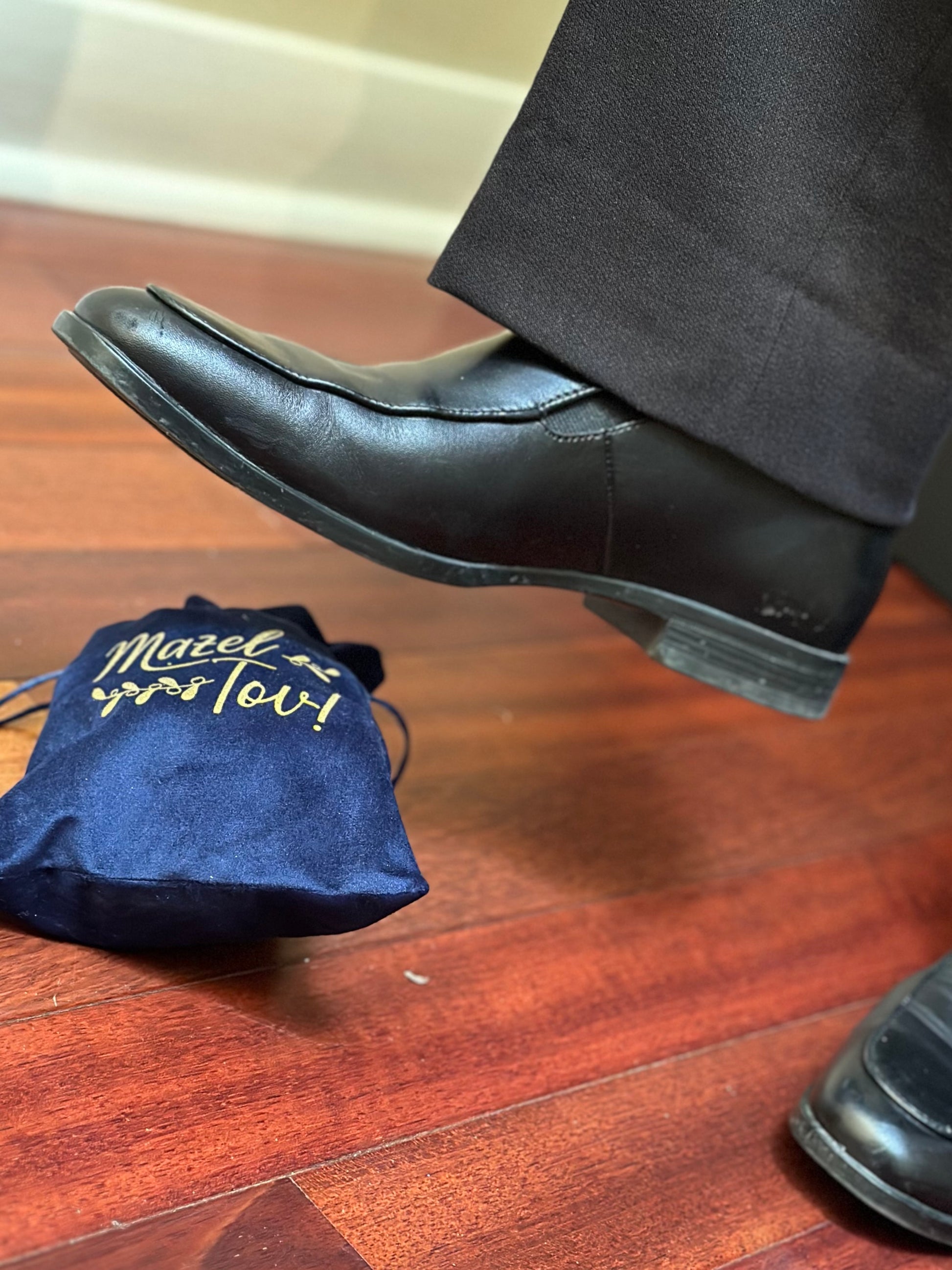 A man's foot stomping on a blue velvet pouch with 'Mazel Tov!' printed on it, containing a Chuppah glass, symbolizing the traditional glass-breaking ritual at a Jewish wedding